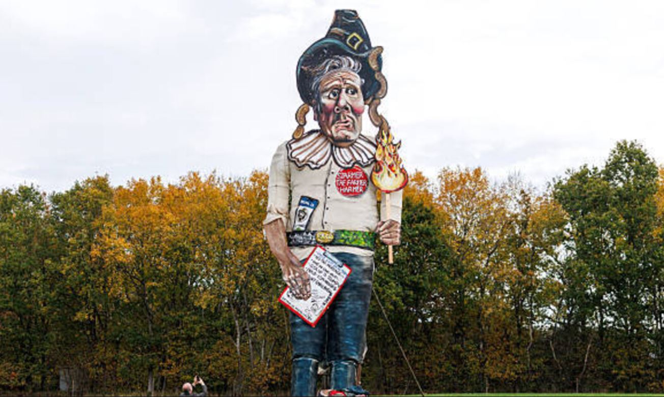 Lutka s podobo britanskega premierja je visoka 11 metrov, nosi klobuk, japonke in gorečo baklo z napisom »Prilizujem se Trumpu«. Vir: Posnetek zaslona, X